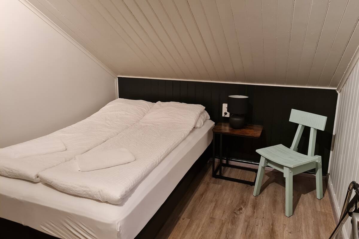 A single bed with a white mattress and pillows, a bedside table with a lamp, and a light blue chair.