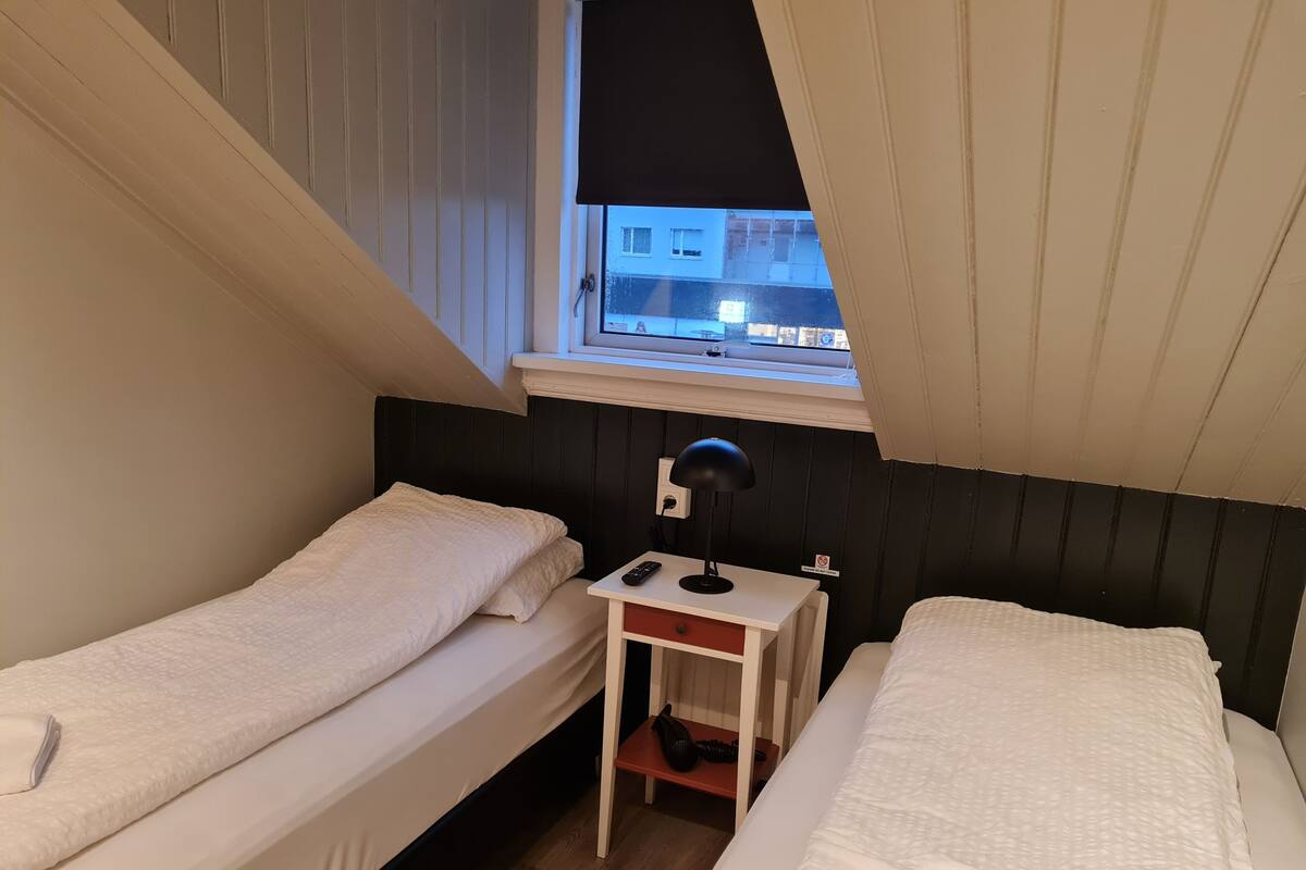 A small, compact room with two single beds, a desk with a lamp, and a window overlooking a building.
