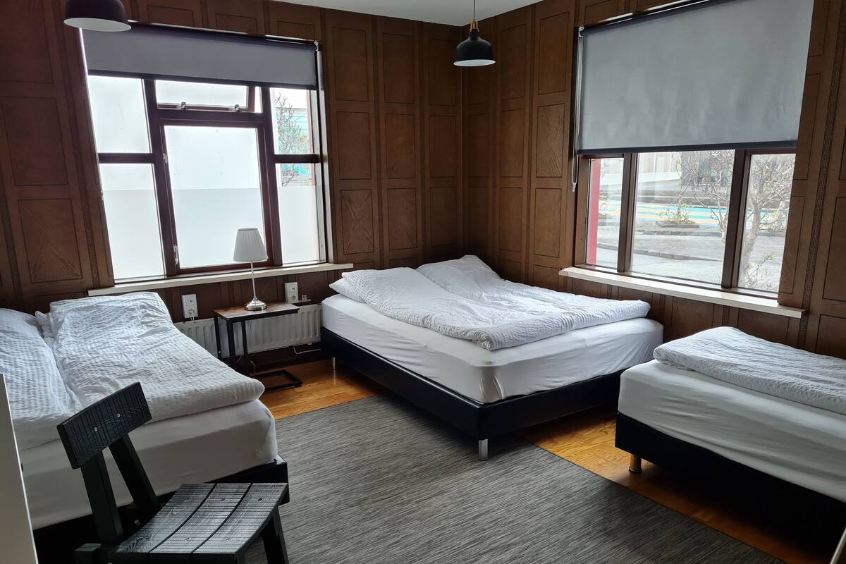 A room with three beds, wooden paneling, a radiator, a small table with a lamp, and large windows with blinds.