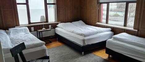 A room with three beds, wooden paneling, a radiator, a small table with a lamp, and large windows with blinds.