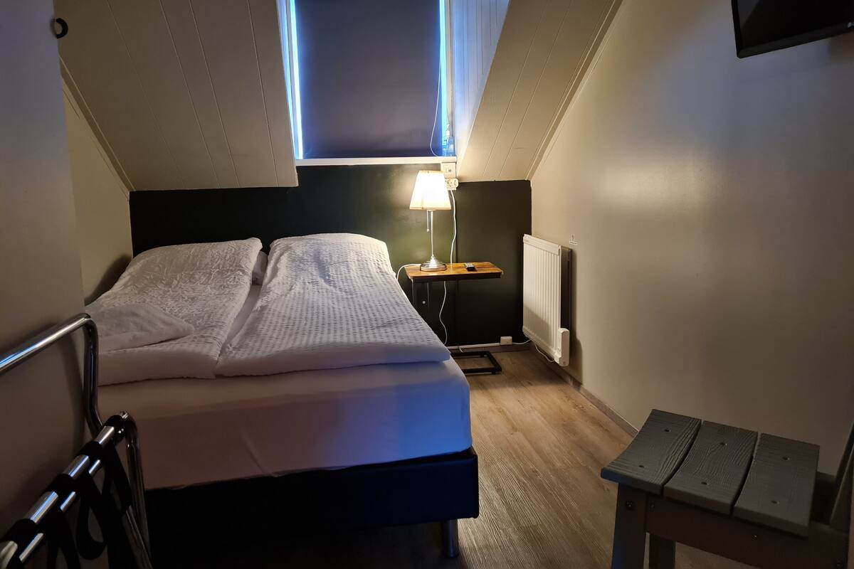 A bedroom with a bed, a bedside table with a lamp, a small table, a radiator, and a window with a view of the sky.