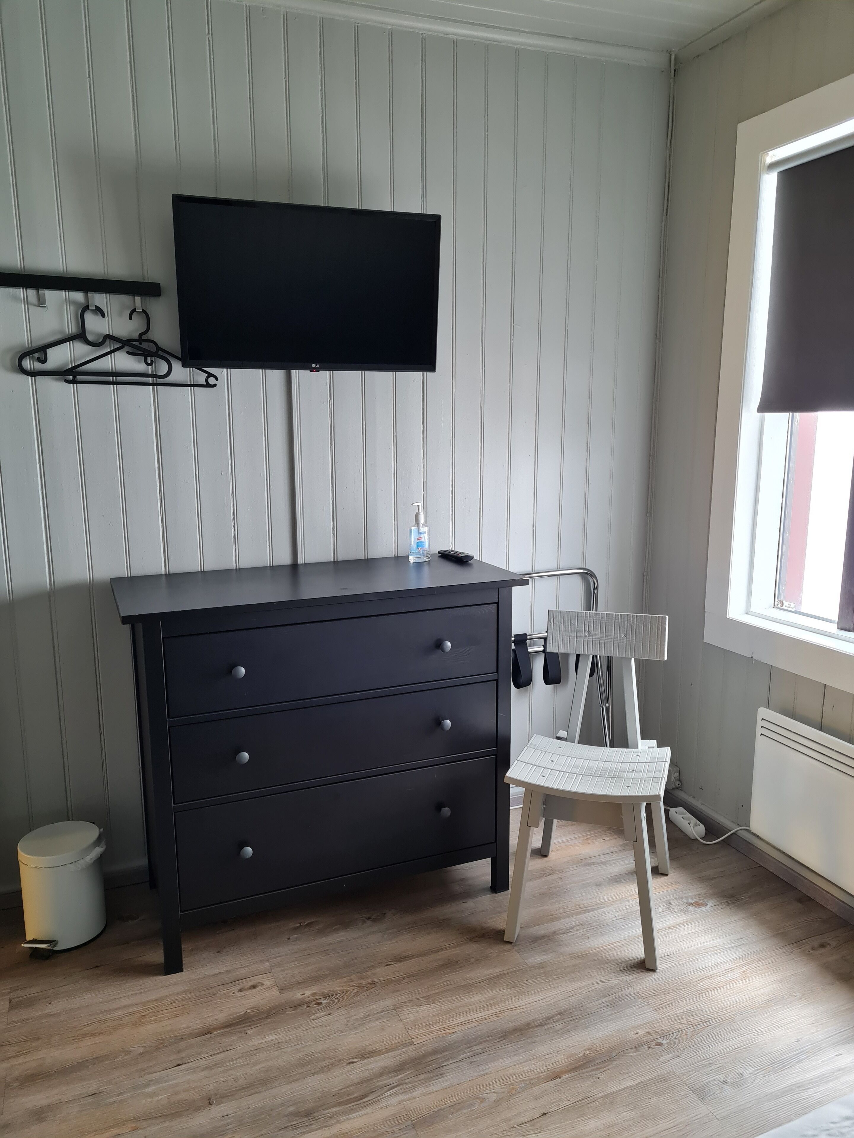 Een kamer met een donkere houten dressoir, een aan de muur bevestigde televisie, een stoel en een raam met zonwering.