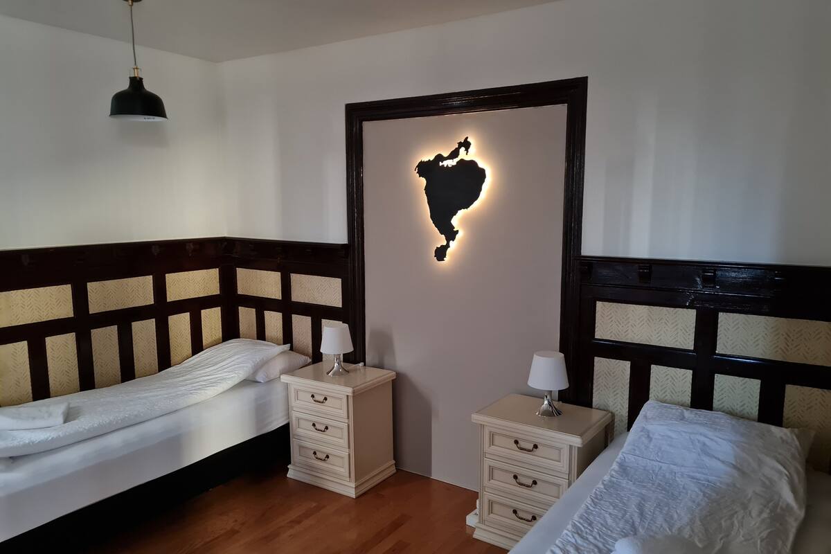 A hotel room with two beds, wooden headboards, a nightstand with drawers, a lamp, and a wall-mounted light fixture.