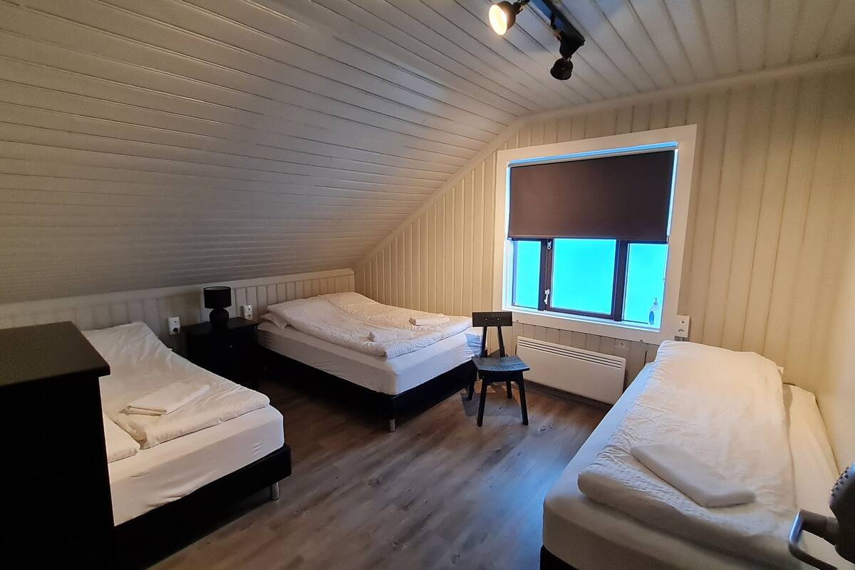 A small, cozy attic bedroom with two beds, a heater, and a window with blinds.