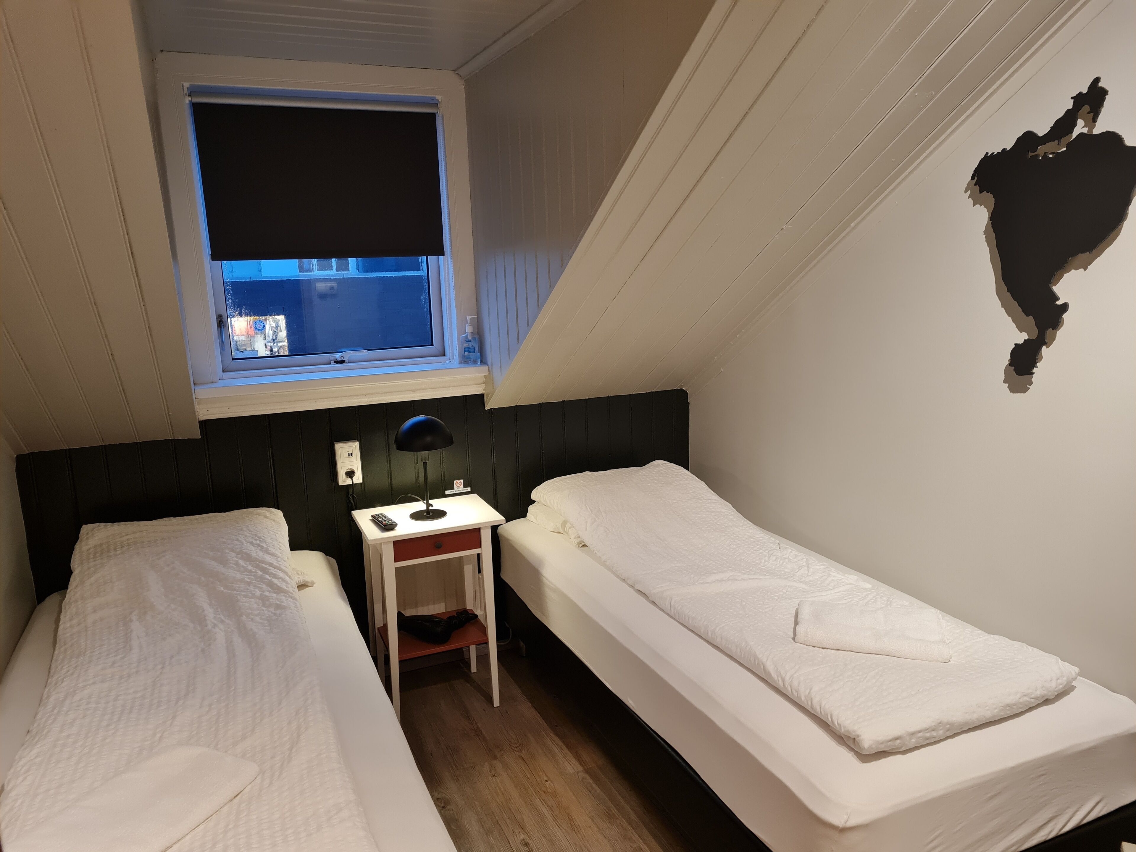 A small, compact room with two single beds, a small table, a lamp, and a window with a view of the street.