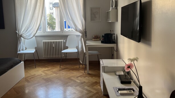 A modern, well-lit room with a bed, a desk, a chair, a radiator, a TV, and a window with curtains. - Cicerone 60 (Rome)