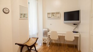 A small, well-lit room with a wooden floor, a desk with a computer and a flat-screen TV, a wicker chair, and a small shelf with decorative items. - Cicerone 60 (Rome)