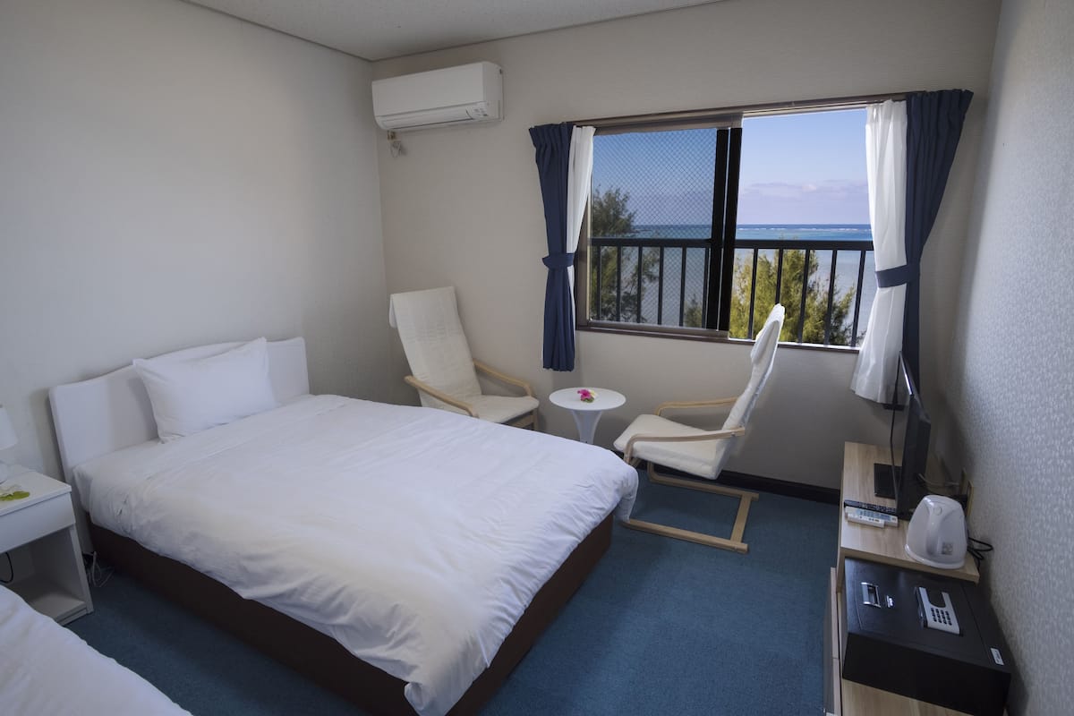 A hotel room with a large bed, two chairs, a small table, a desk with a phone and a kettle, and a window with curtains.