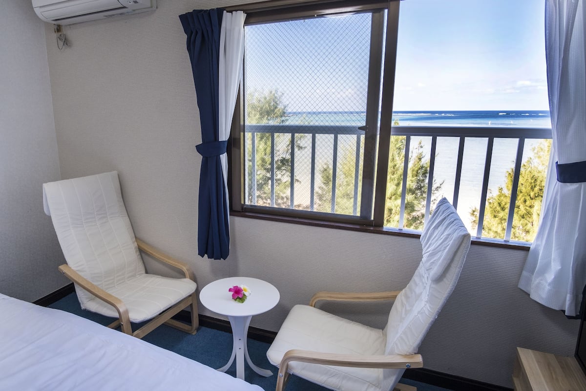 A hotel room with two white upholstered chairs, a small round table with a flower, and a bed. A window with a view of the ocean and trees is visible.