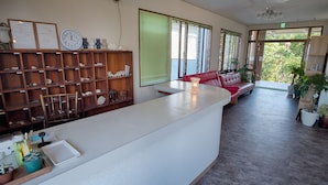 Un área de recepción con mostrador blanco, estanterías de madera con diversos objetos, un sofá rojo y plantas en macetas.