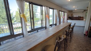 Zona de bar con mostrador de madera y varios taburetes, ventanas amplias con cortinas y vistas al exterior.