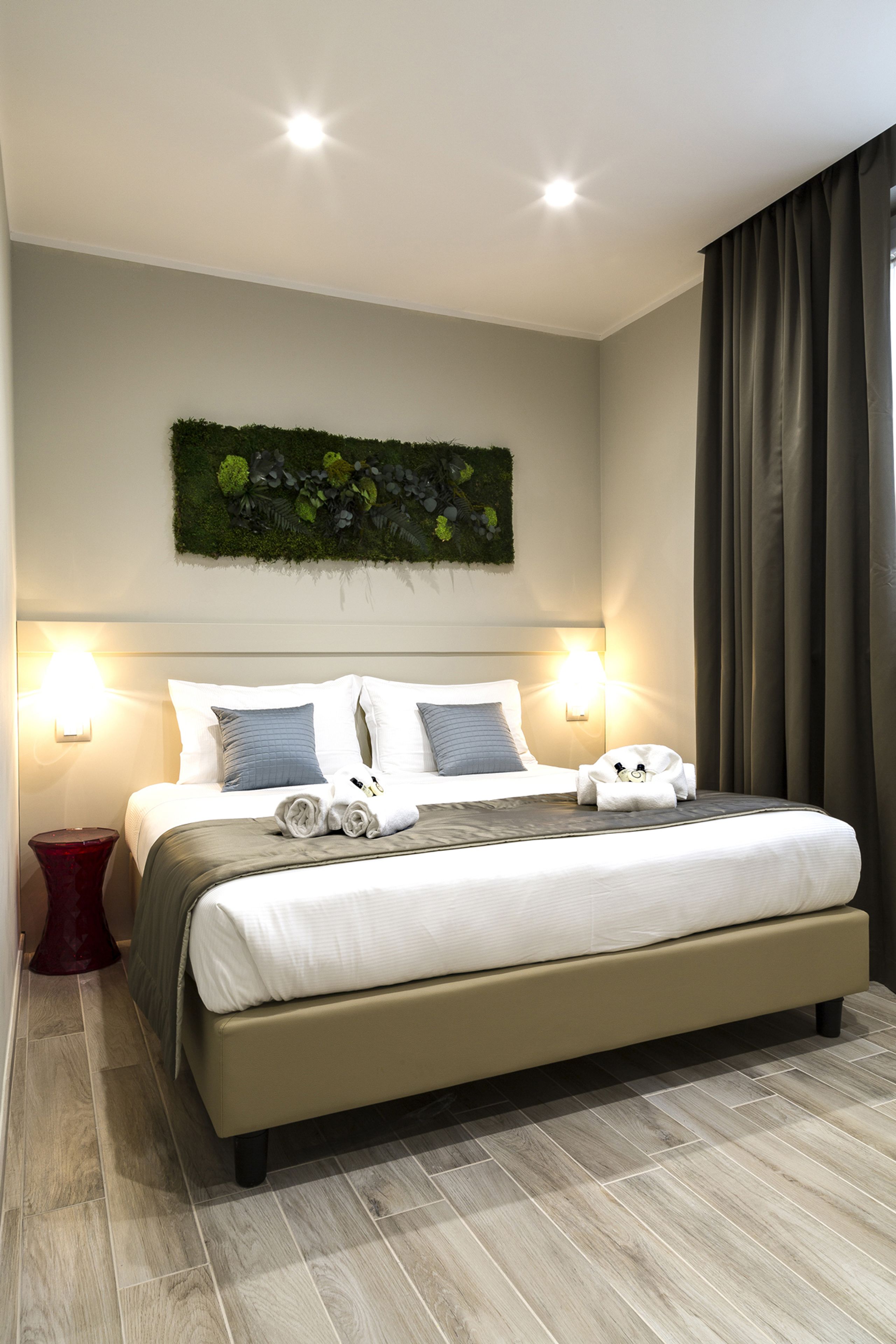 Habitación de hotel moderna con una cama grande, dos lámparas de noche, una mesita de noche roja, un adorno de pared verde y vistas al exterior a través de una ventana con cortinas.