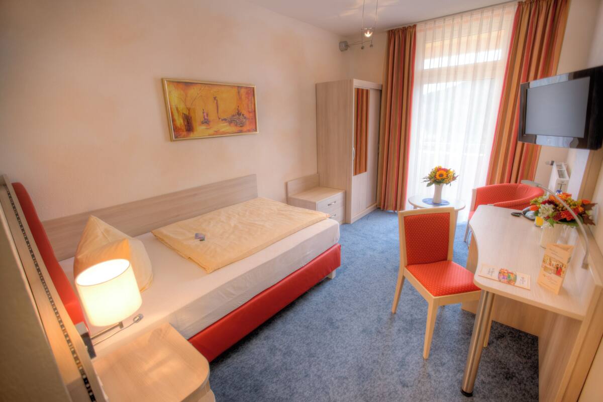 A hotel room with a bed, a desk with a chair, a TV, a painting on the wall, and a window with curtains.