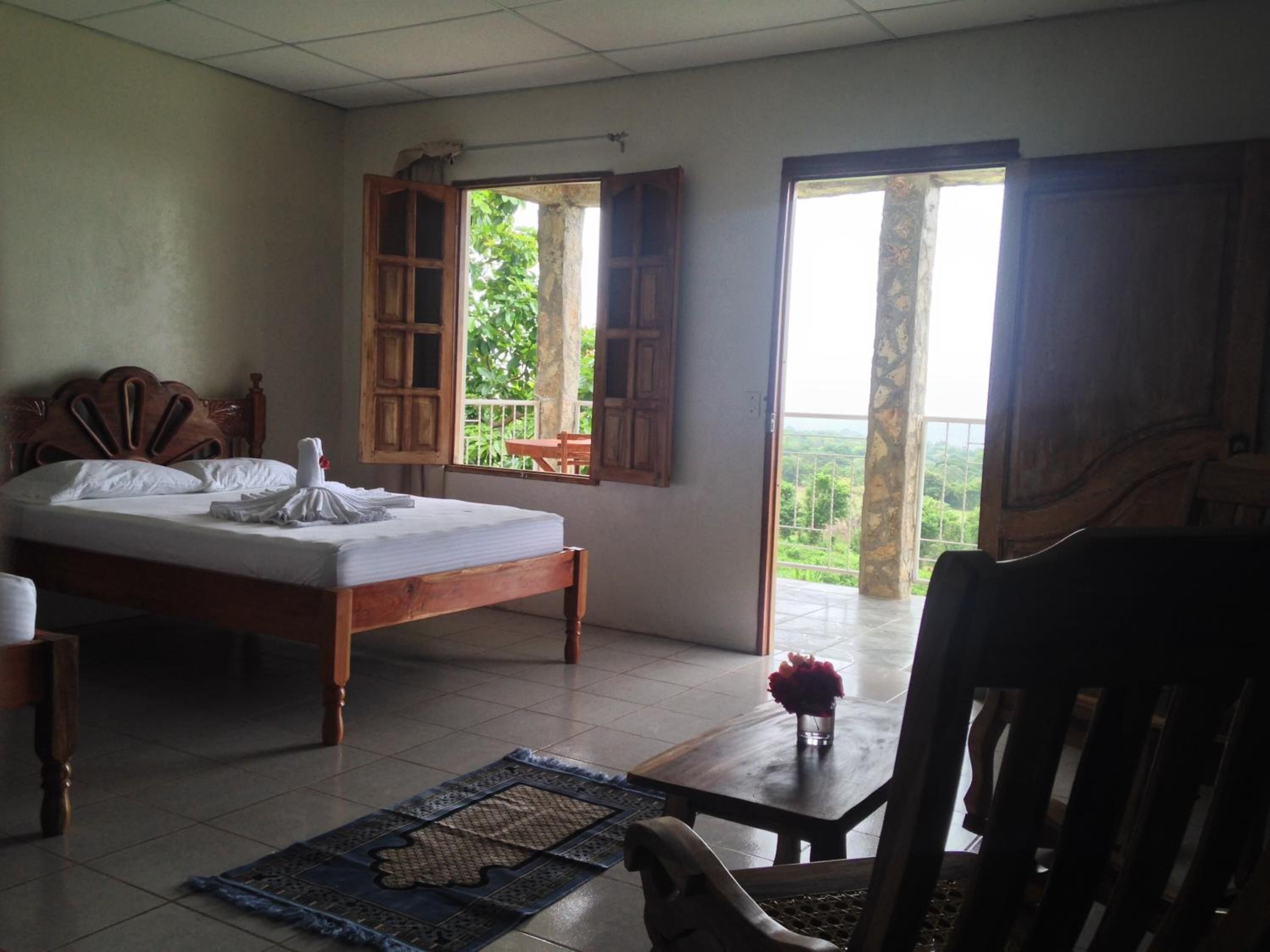 Ein Zimmer mit einem Bett, einem Holzstuhl, einem kleinen Tisch mit Blumenvase und Blick ins Grüne durch offene Fenster.