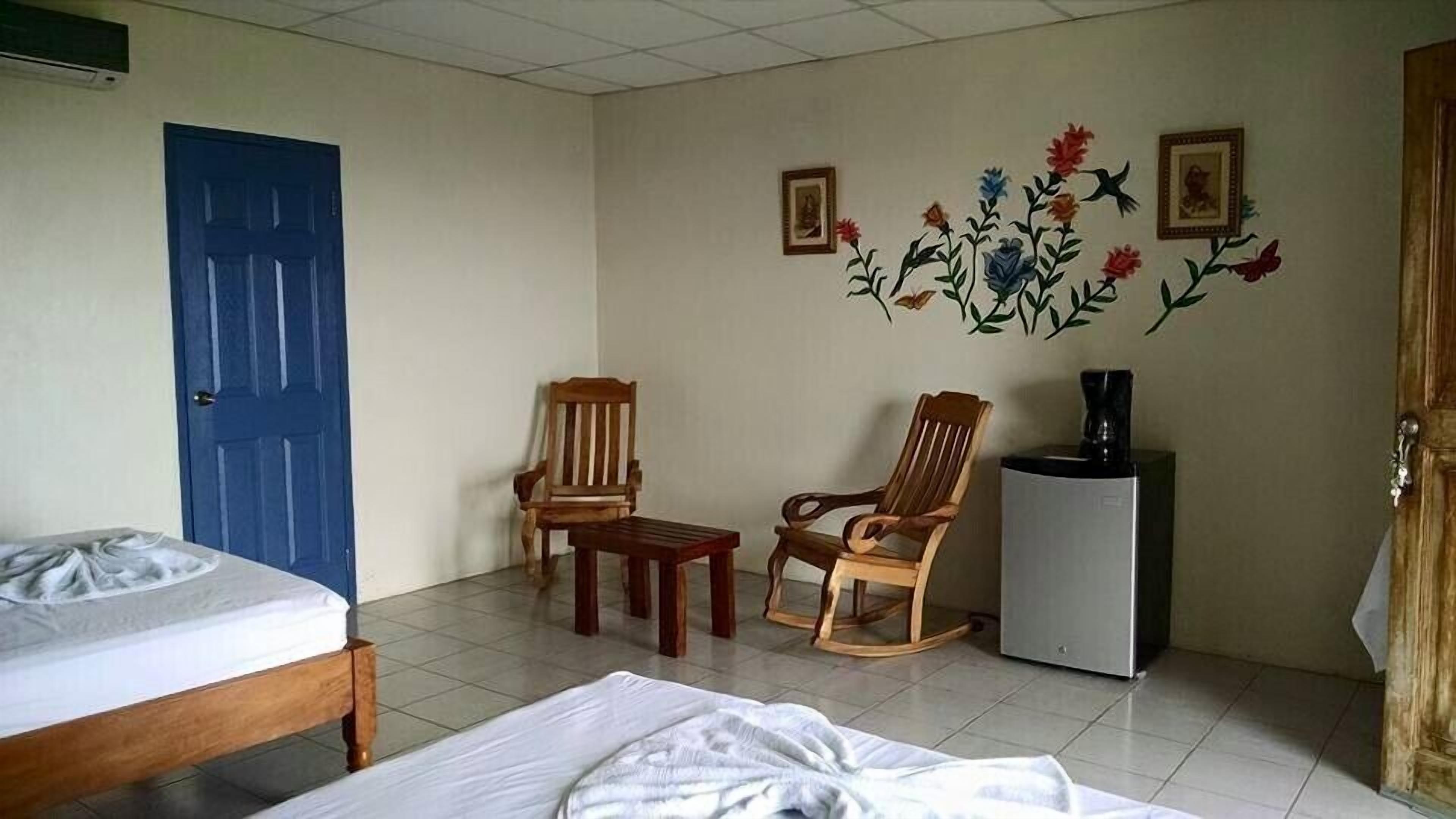 Quarto com uma cama, duas cadeiras de madeira, uma mesinha, um refrigerador e uma porta azul.