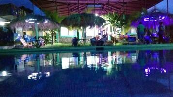 A pool area with a thatched-roof structure, lounge chairs, and a group of people gathered around a table.