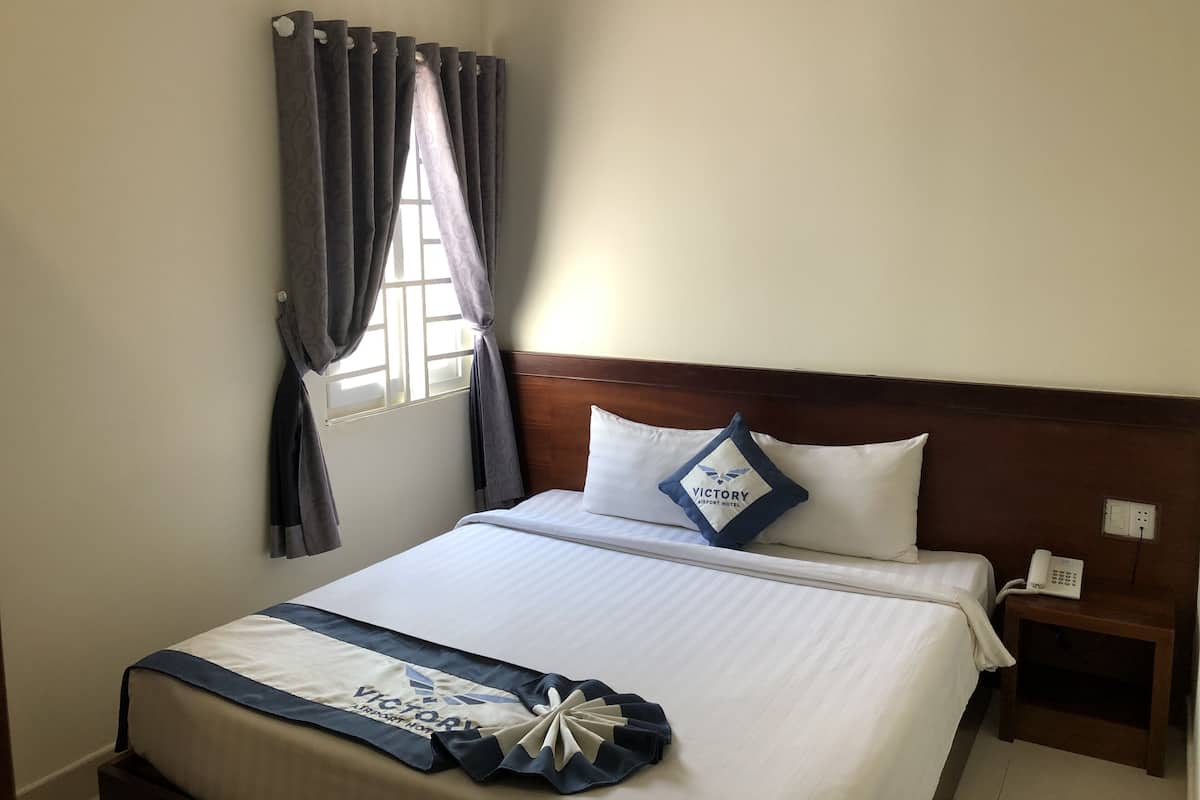 A neatly made bed with a white and blue bedspread, a pillow with "VICTORY AIRPORT HOTEL" text, and a bedside table with a phone.
