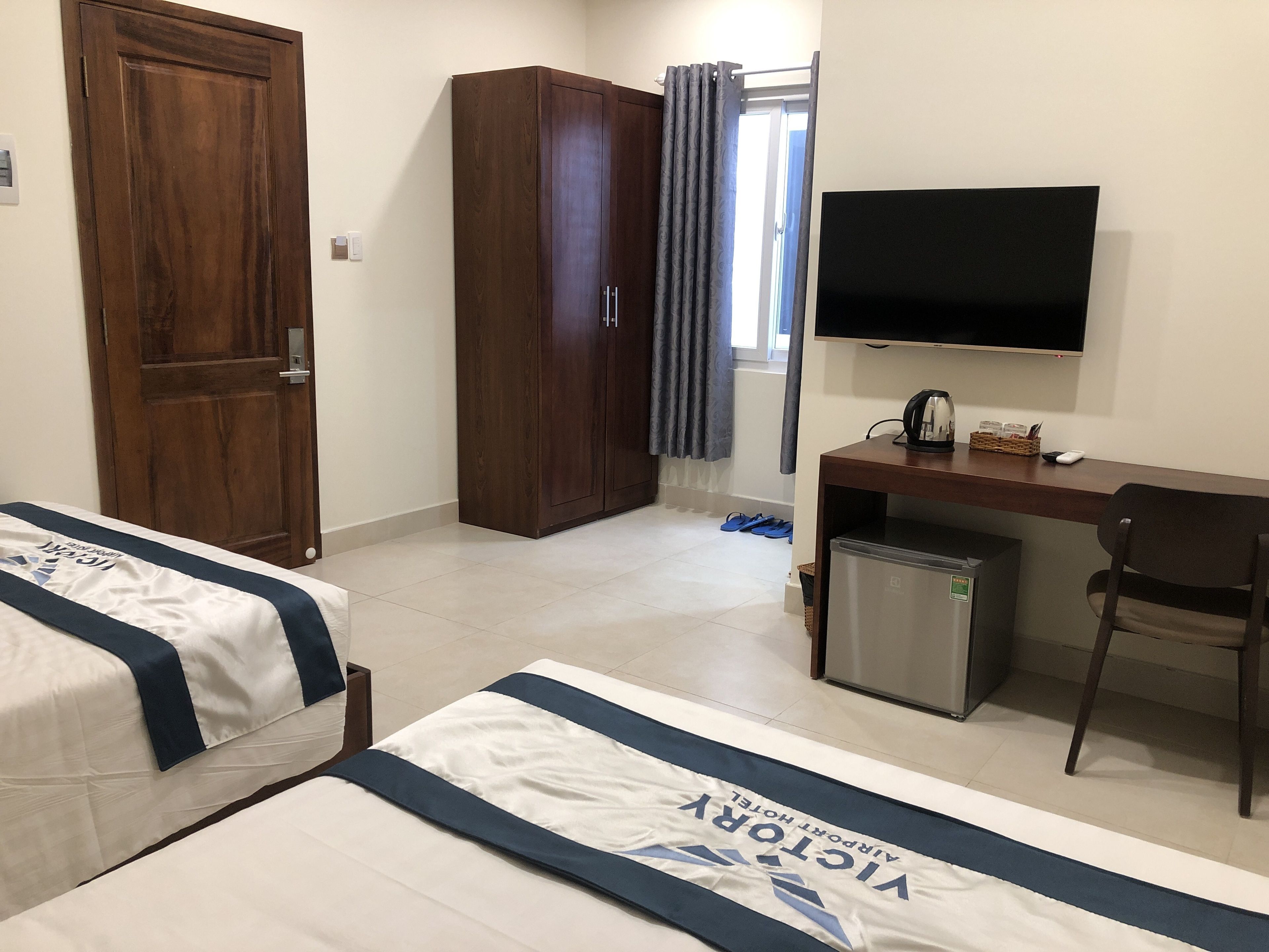 A hotel room with two beds, a desk with a TV, a wardrobe, and a small fridge.