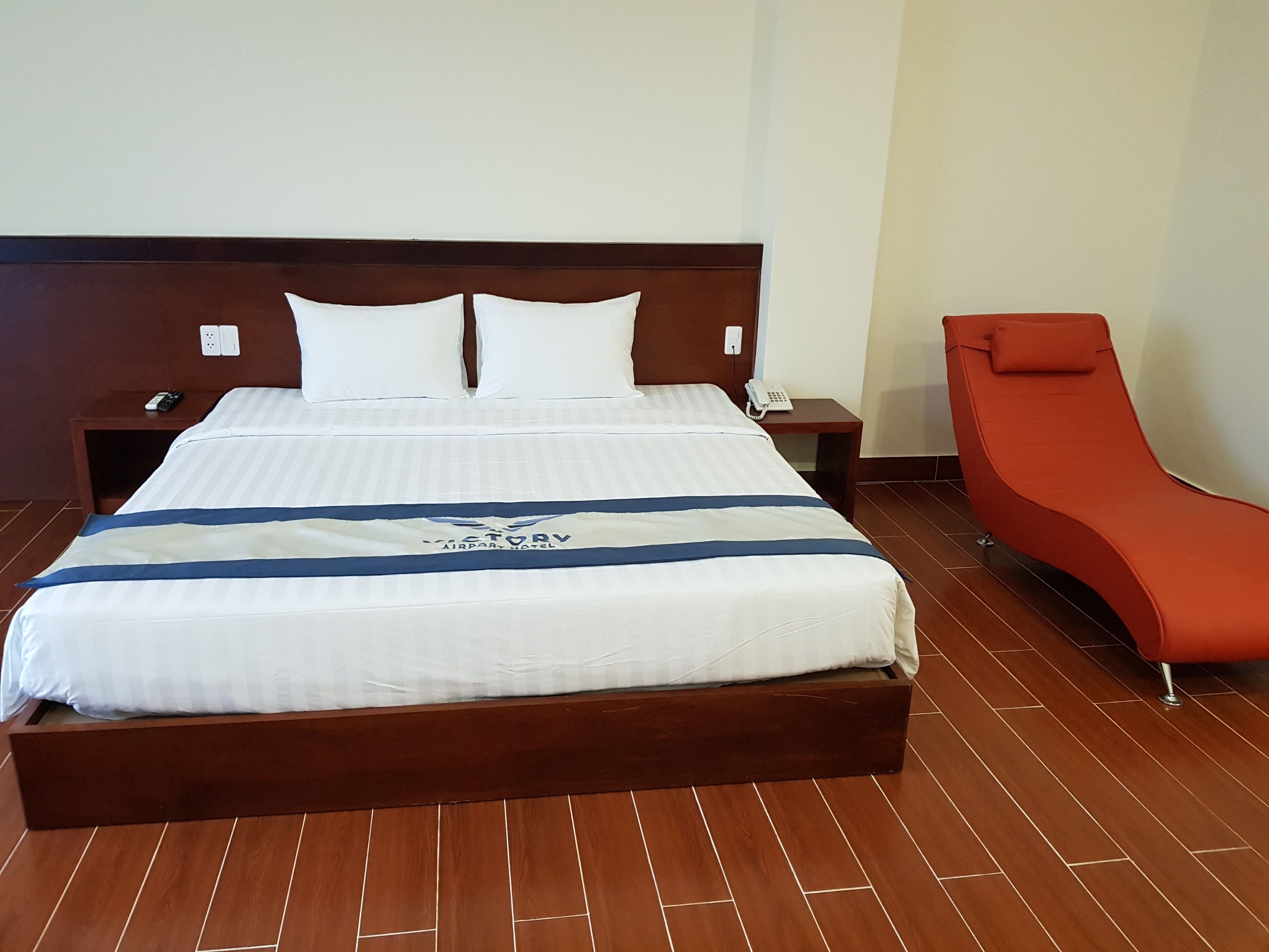 Quarto de hotel com uma cama grande, mesa de cabeceira com telefone, poltrona vermelha e piso de madeira.