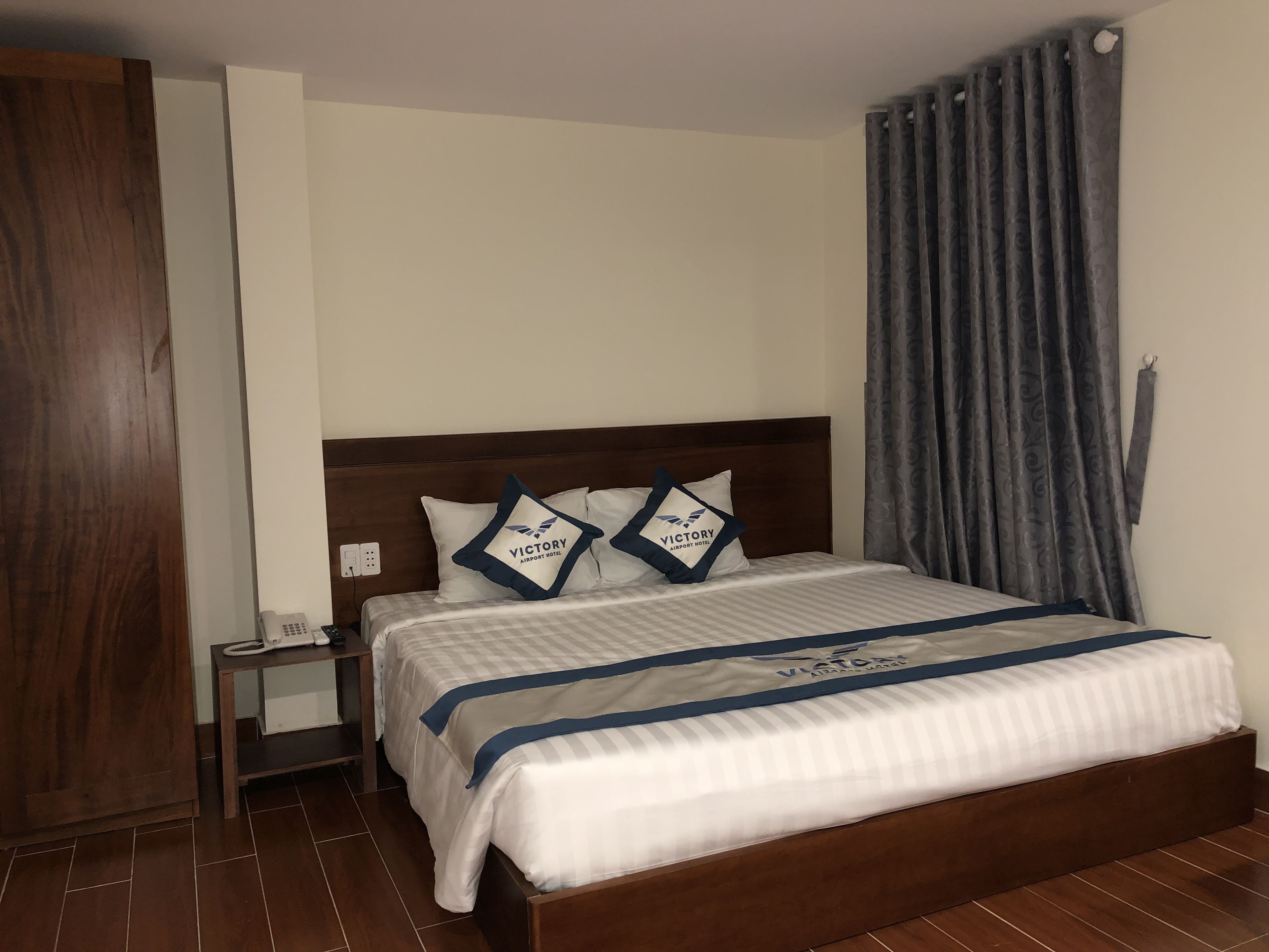 Een hotelkamer met een houten hoofdeinde, een bed met wit beddengoed en blauwe accenten, een nachtkastje met een telefoon en een grijze gordijn.