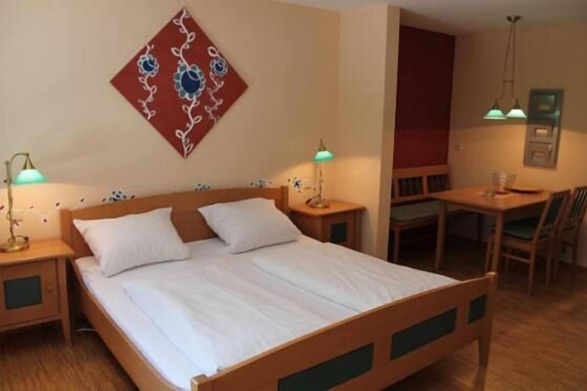 Un lit simple avec du linge de lit blanc, une tête de lit en bois, deux tables de chevet avec des lampes, une petite table avec des chaises et une œuvre d’art accrochée au mur.