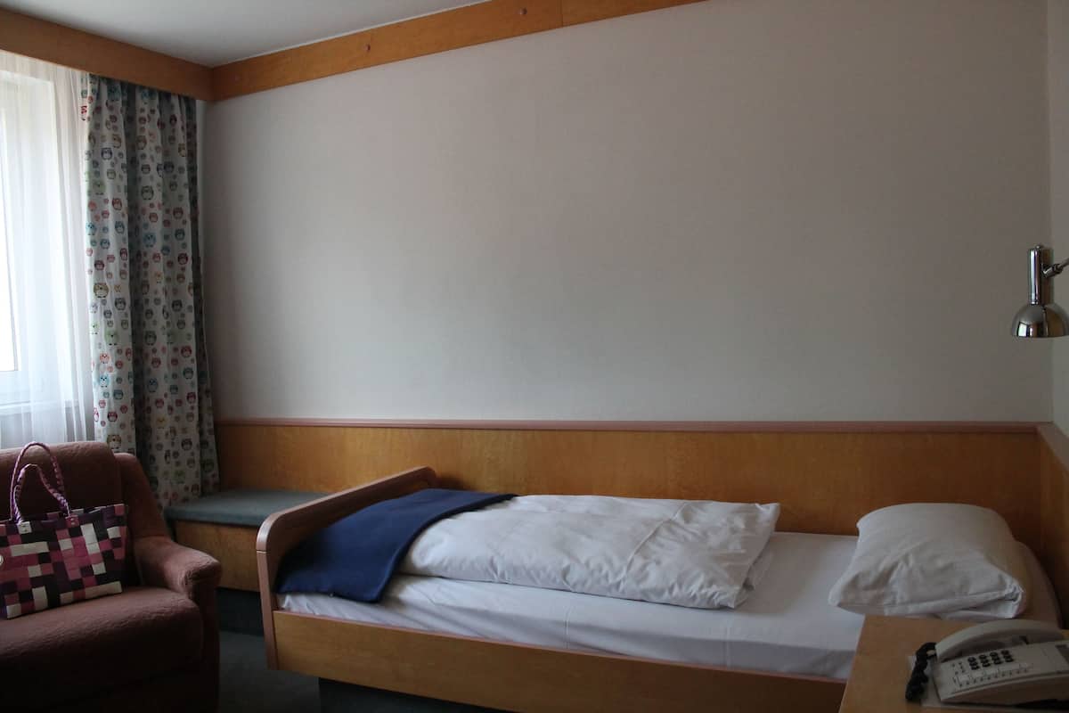 Une chambre d’hôtel avec un lit simple, une tête de lit en bois, une table de chevet avec un téléphone et une chaise avec un sac à motifs.