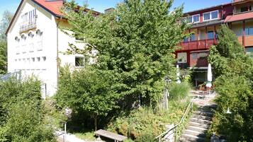 En bygning med flere etager og rødt tag, omgivet af frodig vegetation og en stentrappe op til en træbænk.