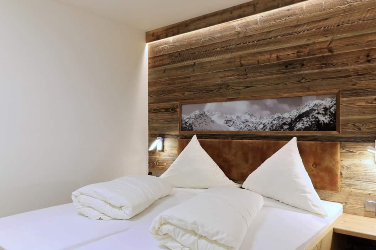 Een netjes opgemaakt bed met witte lakens en kussens, een houten hoofdbord en een lijst met een afbeelding van een besneeuwd berglandschap.
