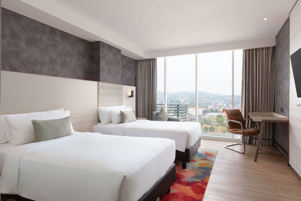 Quarto de hotel moderno com duas camas, uma mesa de trabalho e uma grande janela com vista para a cidade.