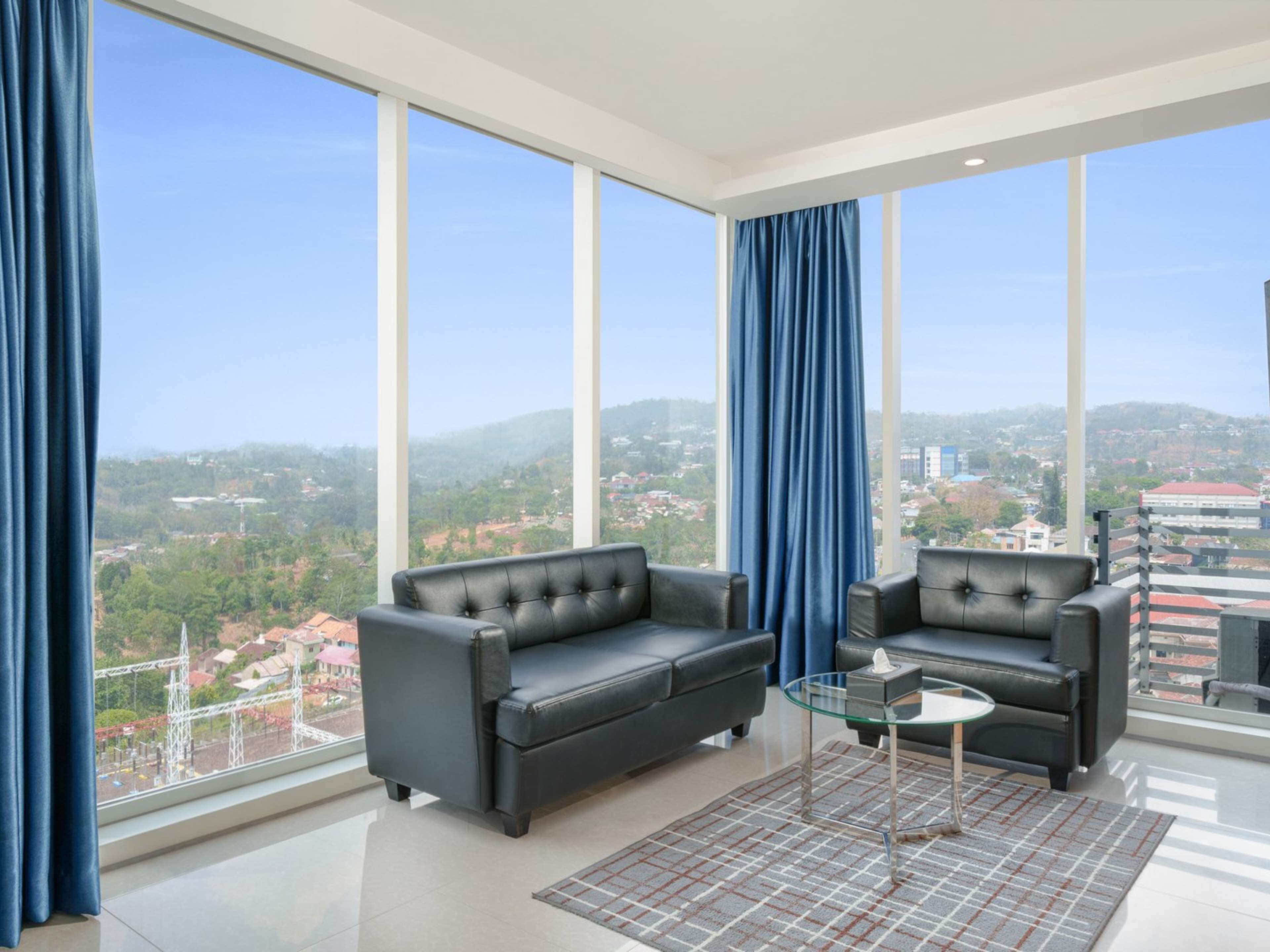 Una sala moderna con un ventanal grande, dos sofás de cuero negro, una mesa de centro de vidrio y vistas a un paisaje urbano.