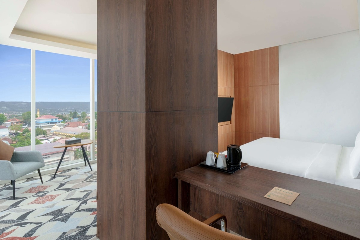 Quarto de hotel com uma janela grande, uma escrivaninha de madeira, uma cama e uma cadeira.