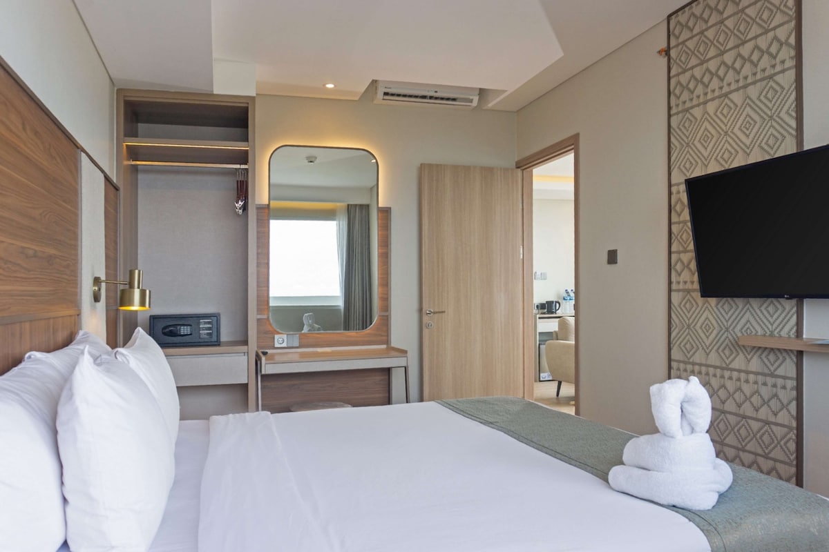 Quarto de hotel com cama, mesa de cabeceira, espelho, televisão e banheiro visível através de uma porta aberta.