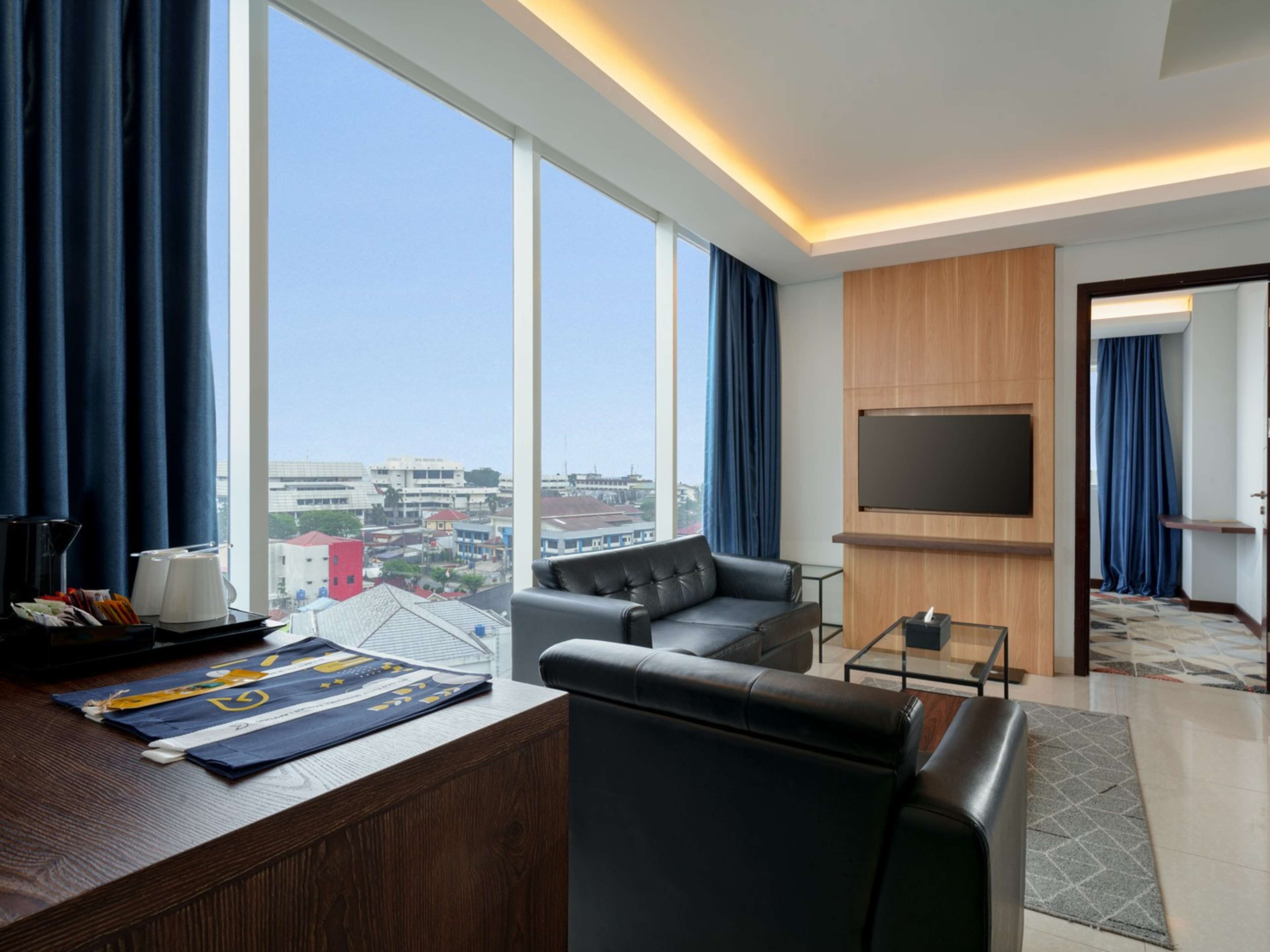 Una habitación de hotel moderna con un ventanal grande, un sofá de cuero, una mesita de centro pequeña y un televisor de pantalla plana montado en la pared.