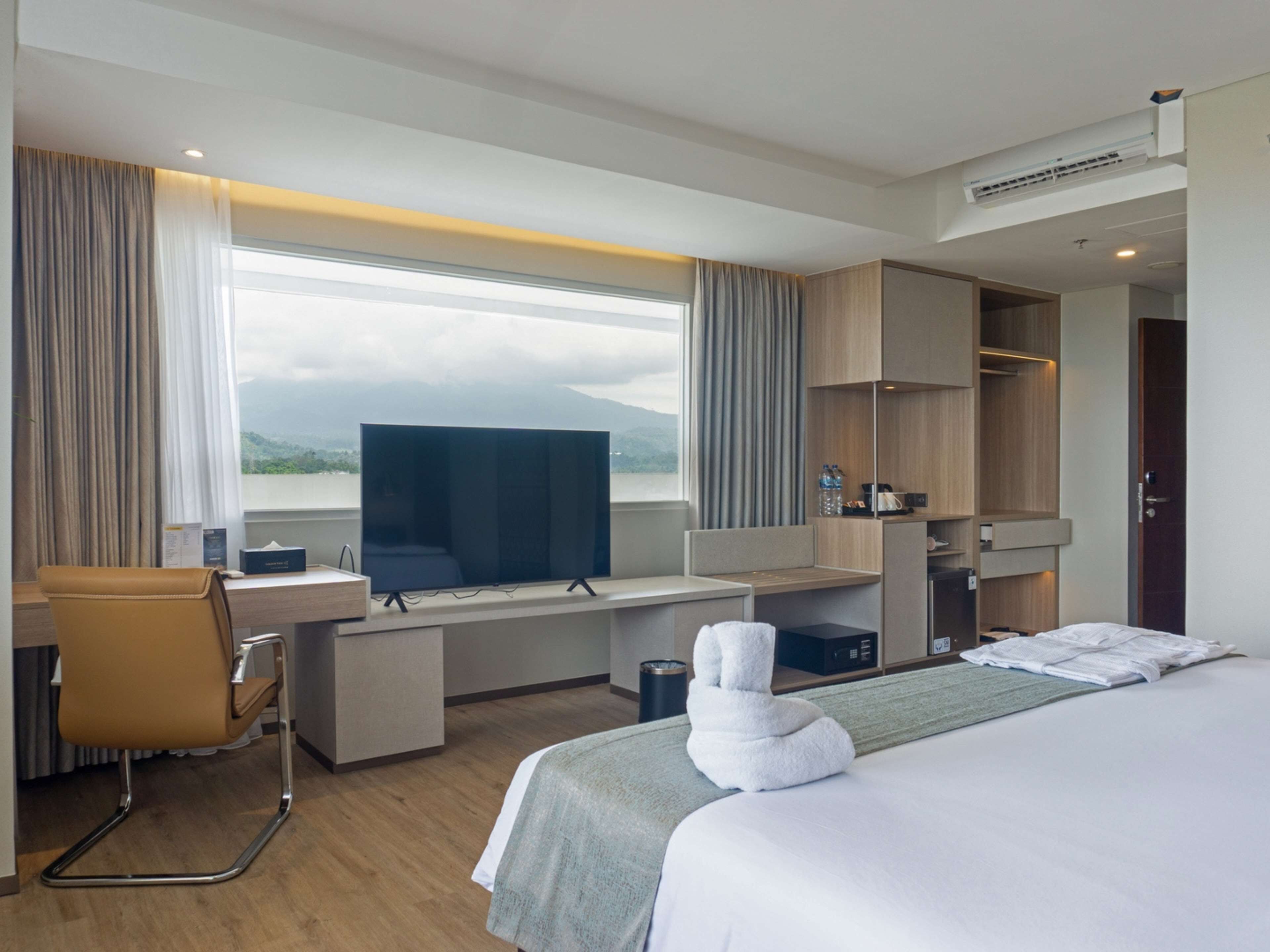 A modern hotel room with a large bed, a desk with a computer, a chair, and a view of mountains through the window.
