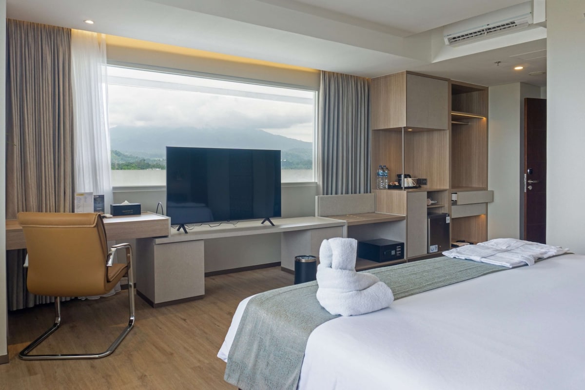 Quarto de hotel moderno com uma cama grande, uma escrivaninha com computador, uma cadeira e vista para montanhas pela janela.