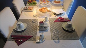 Mesa para cuatro personas dispuesta con sillas blancas, un plato de fruta, pasteles y una estación de café.
