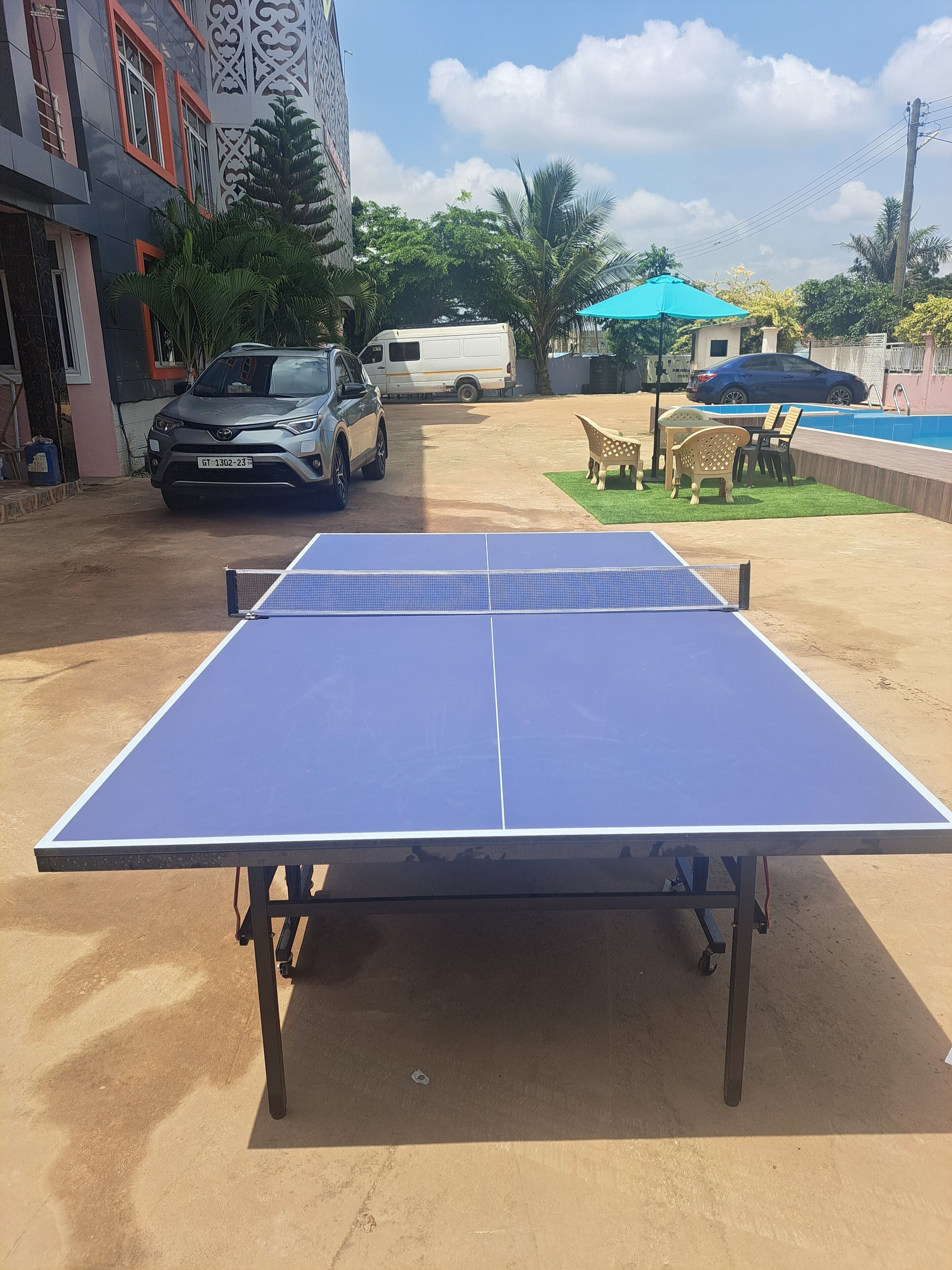 A ping pong table in a courtyard with a car and a van parked nearby, surrounded by greenery and a swimming pool.