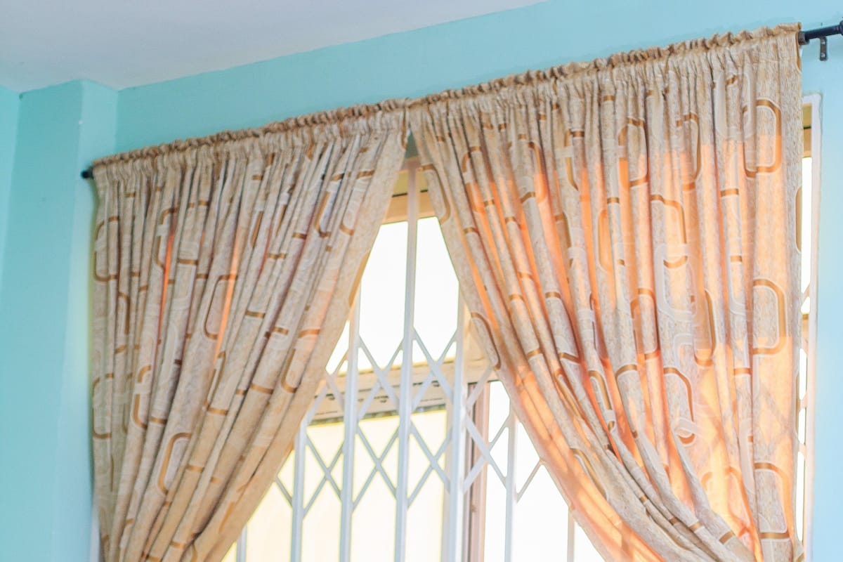 Una ventana con barrotes metálicos blancos y cortinas beige con un diseño geométrico.