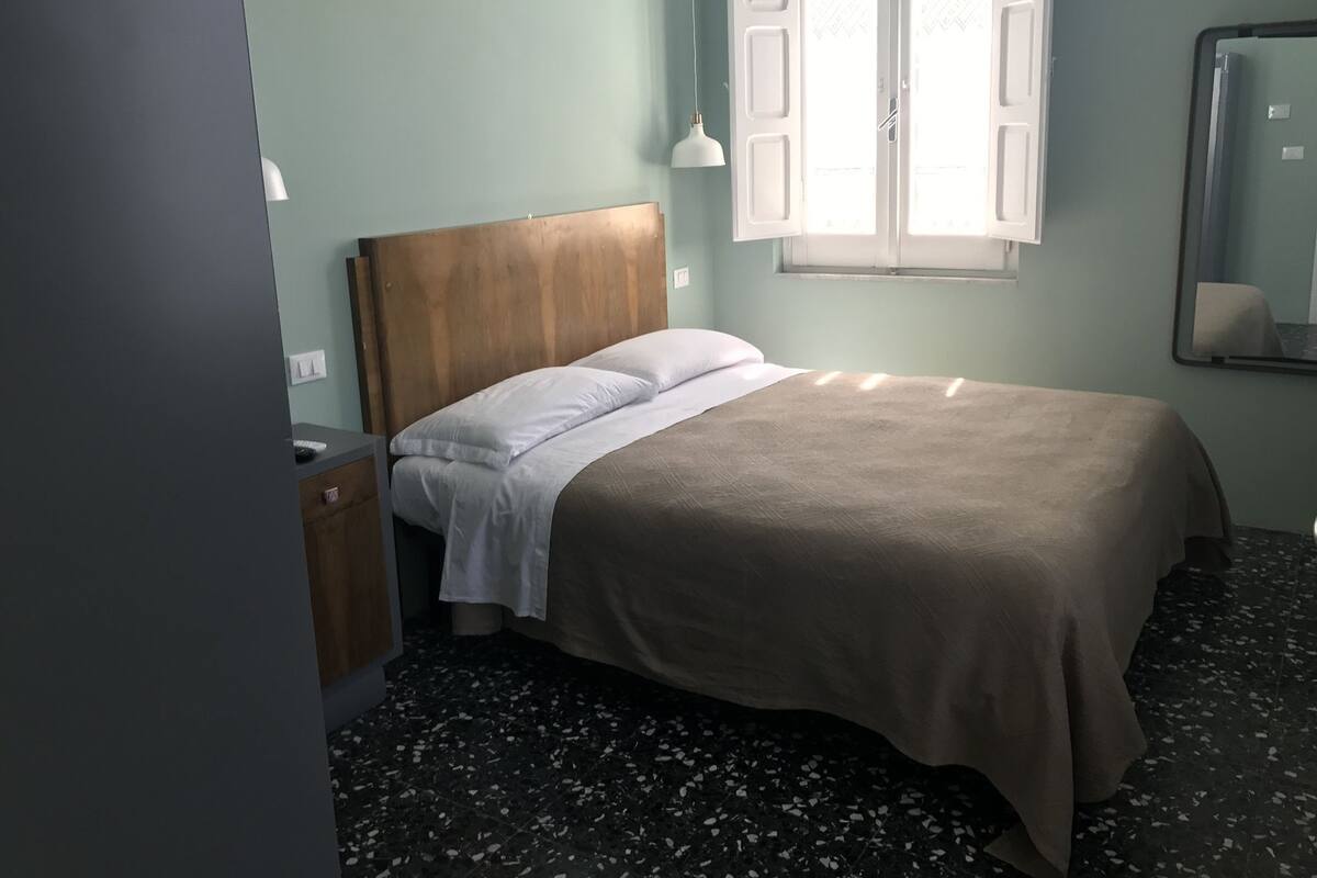 A bedroom with a bed, a nightstand, a mirror, and a window with open shutters.