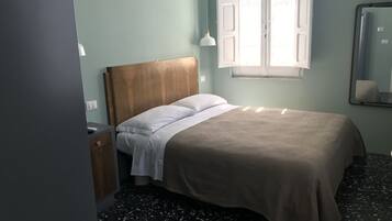 Quarto com cama, mesa de cabeceira, espelho e janela com persianas abertas.
