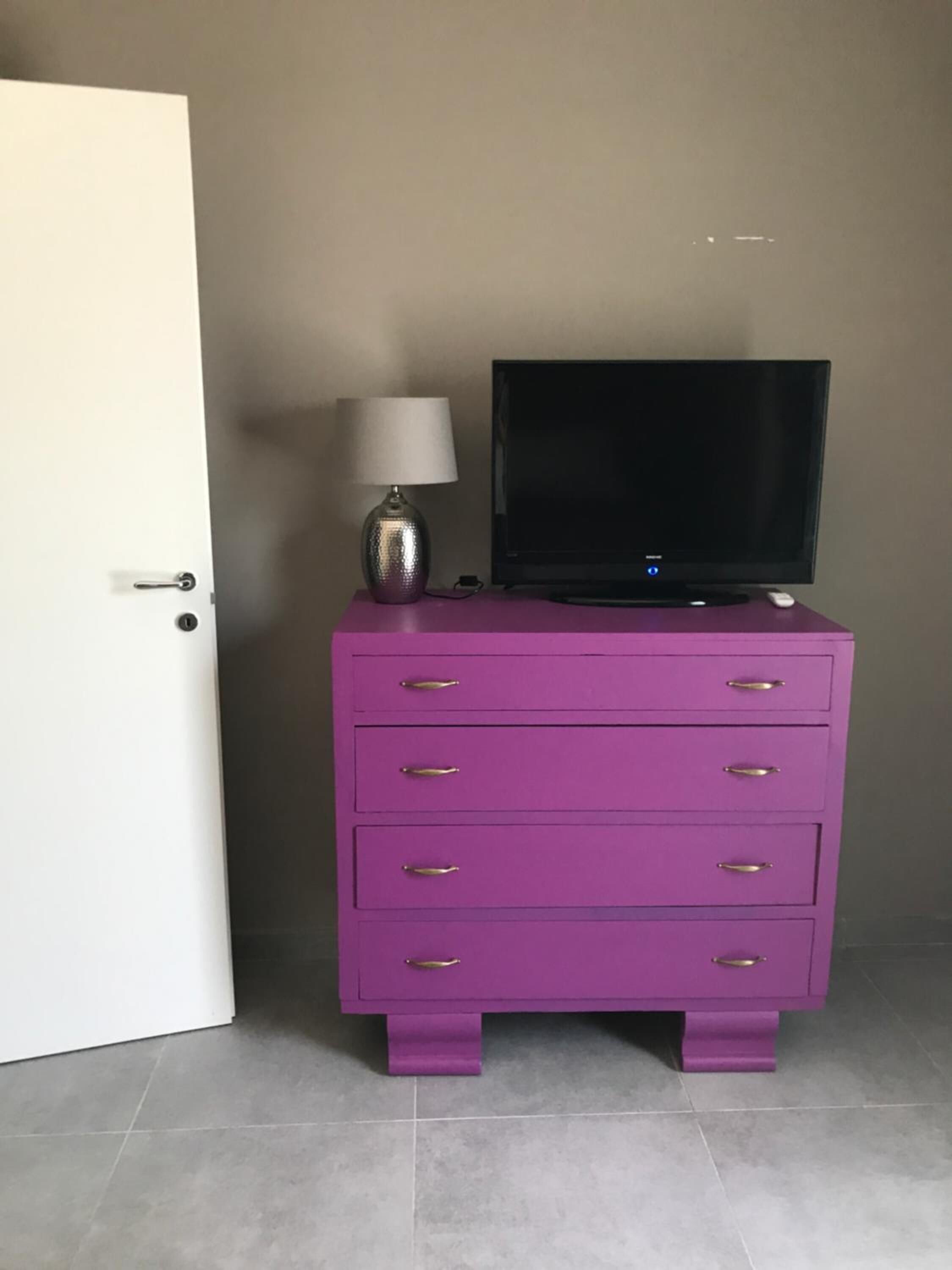 Uma cômoda roxa com uma televisão de tela plana em cima, uma luminária e uma porta à esquerda.