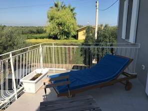 A balcony with a wooden chair and a blue mat, a white pot with a plant, and a view of a yellow house and greenery in the background.
