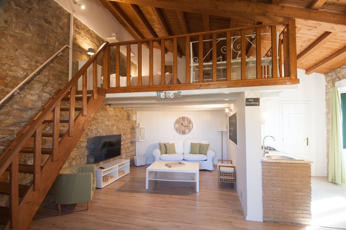 Un espacio habitable amplio con una escalera de madera, un conjunto de sofá blanco, un televisor de pantalla plana y una pared de ladrillo decorativa.