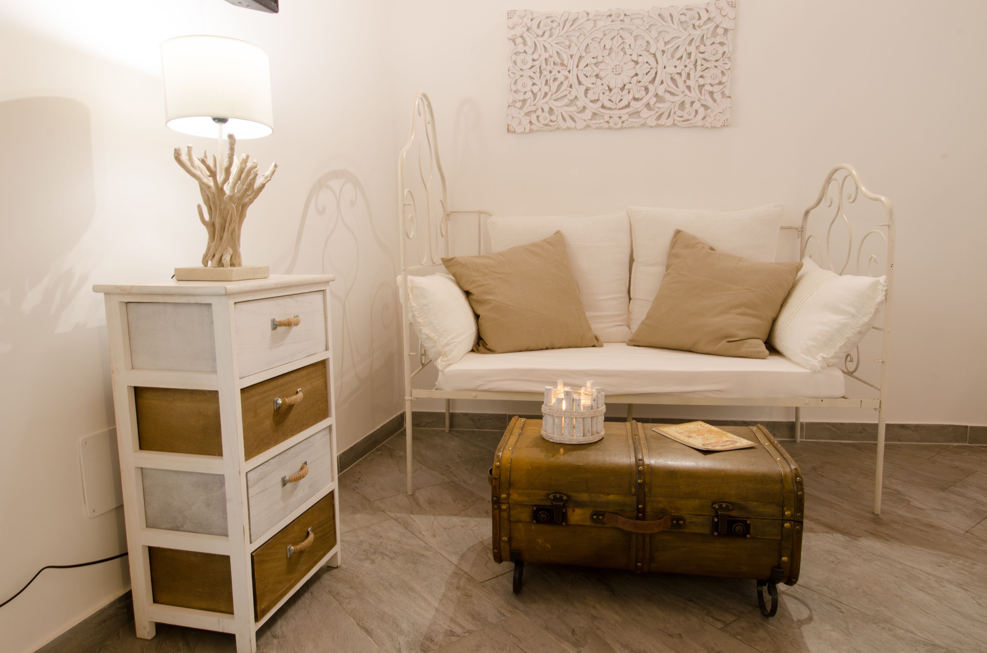 A white sofa with beige cushions, a white bedside table with drawers, a lamp, and a vintage suitcase on the floor.