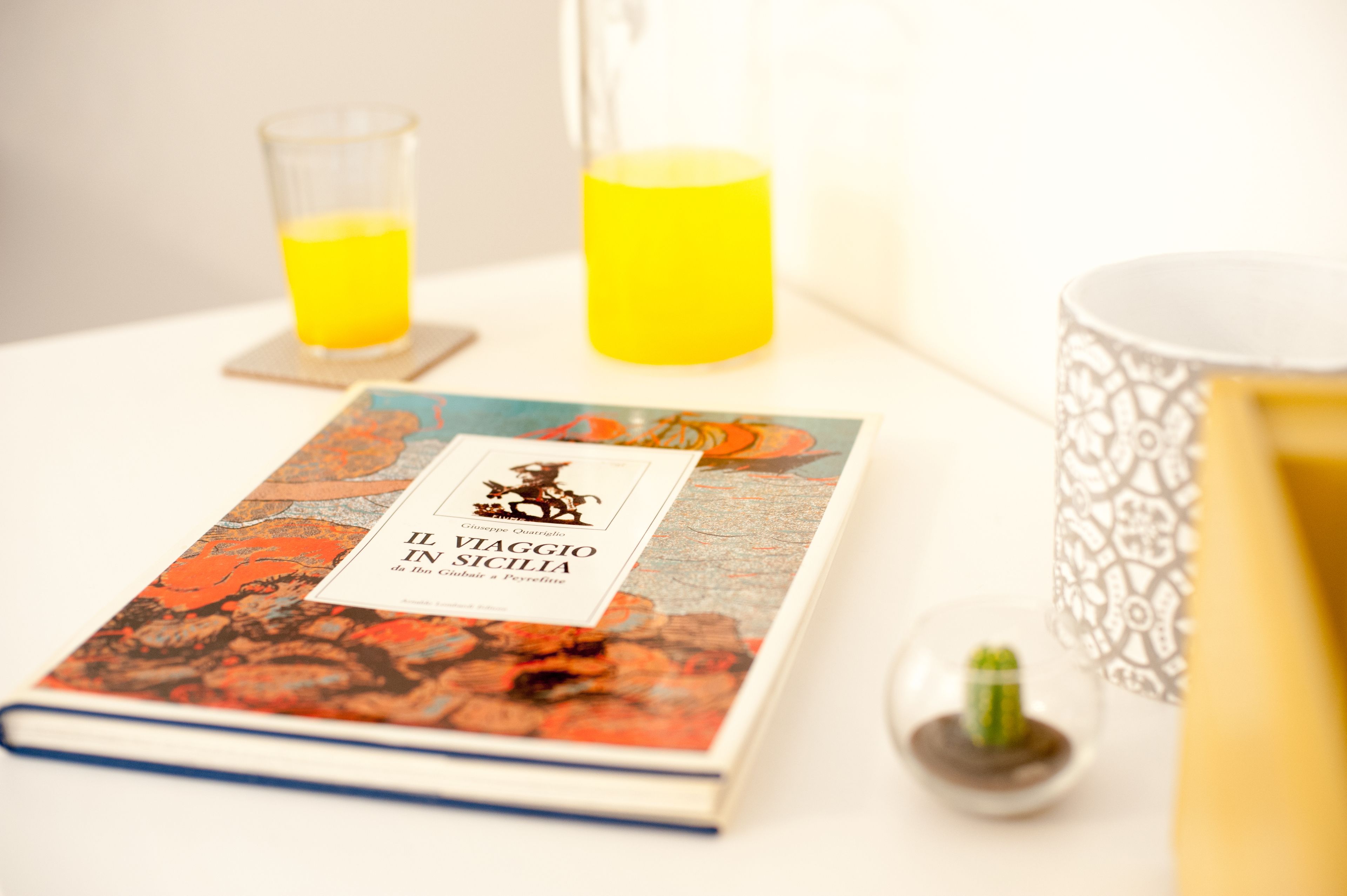 Un livre intitulé « Il Viaggio in Sicilia » est posé sur une table, à côté d’un verre de jus d’orange et d’un récipient en verre décoratif.