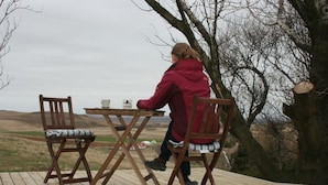 Une personne est assise à une table en bois sur une terrasse extérieure, d’où l’on peut admirer un paysage vallonné.