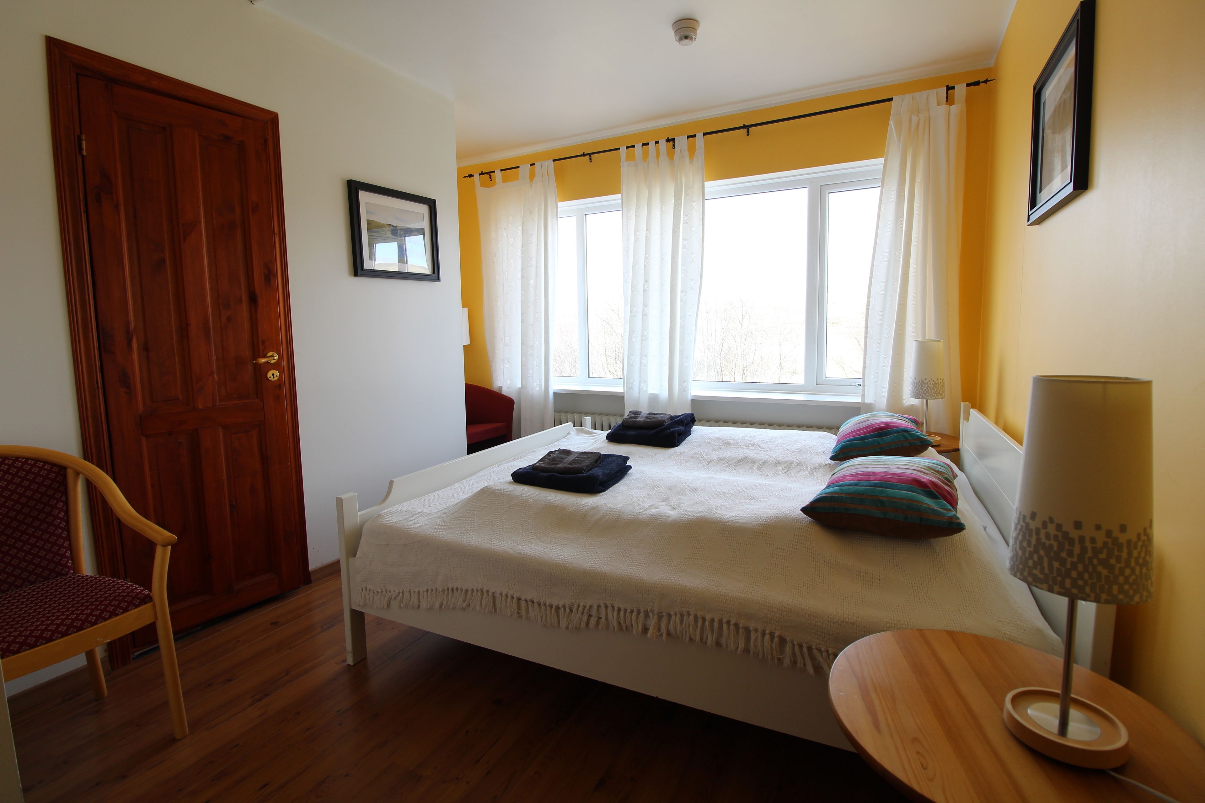 A bedroom with a bed, a chair, a bedside table, a lamp, and a window with curtains.