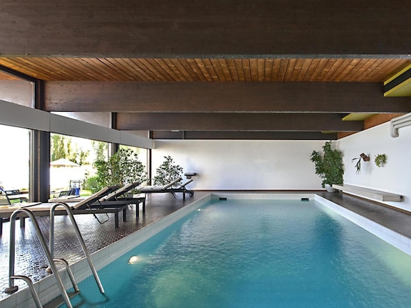 An indoor swimming pool with a wooden ceiling, clear glass walls, and a few lounge chairs.