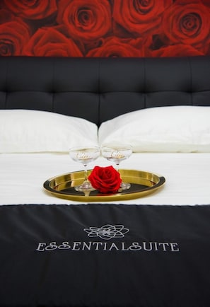 A bed with a black headboard, white pillows, and a tray with two wine glasses and a red rose. The bedspread has "ESSENTIALSUITE" printed on it. - Essential Suite B&B Botanic (Naples)