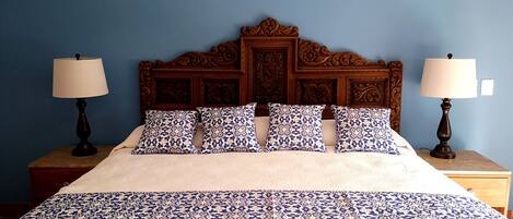 Una cama con una colcha y cojines estampados, flanqueada por dos mesitas de noche con lámparas, frente a una pared azul.